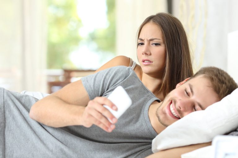 Woman looking over a mans shoulder trying to read his phone while in bed