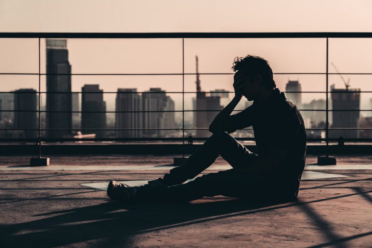Silhouette of a man on a rooftop sitting down while holding his hand to his face in dismay. 