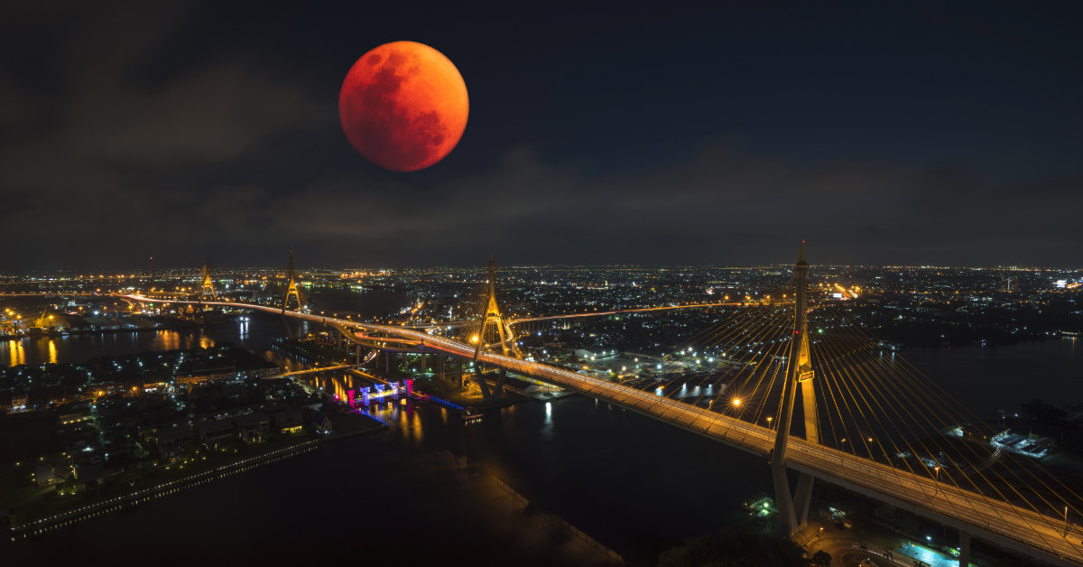 a bloodmoon seen over top of a light filled city, a bridge is seen in the foreground