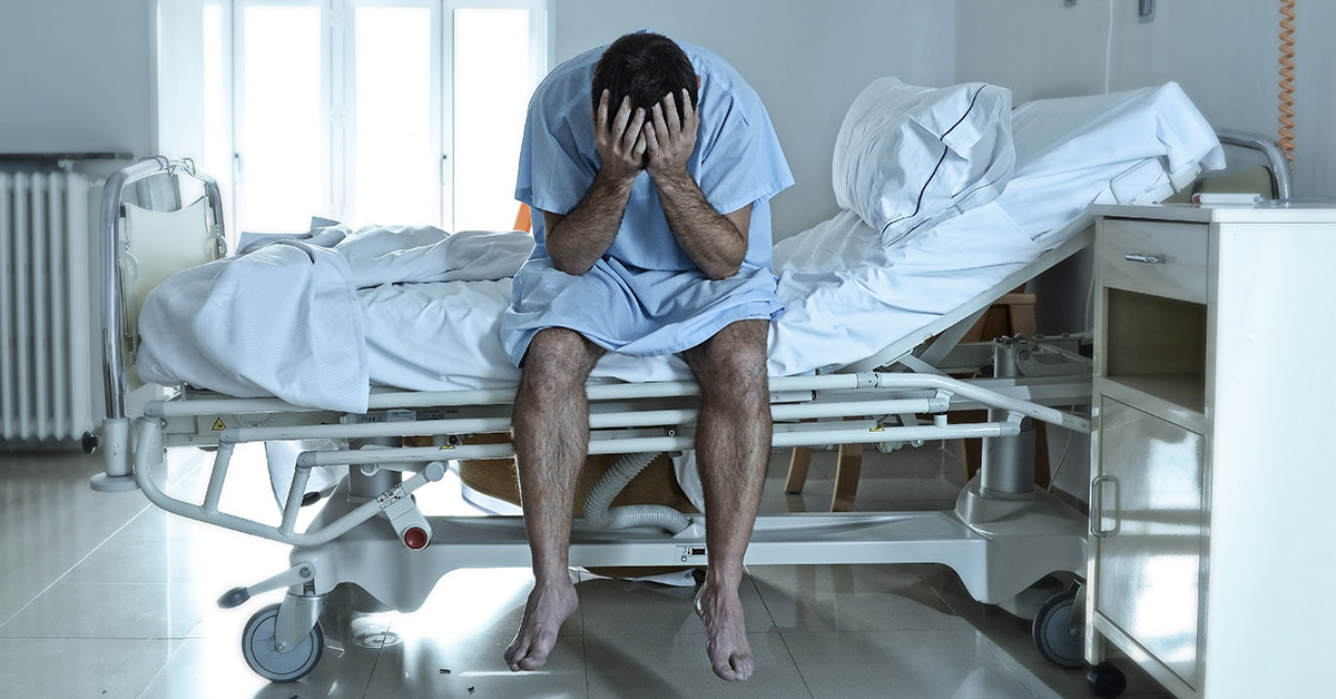 man sitting on edge of hospital bed with with face in palms in despair