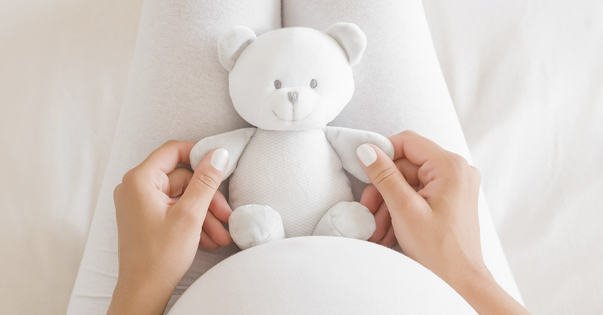 POV: pregnant woman wearing all white looking down holding a white stuffed bear in her lap.