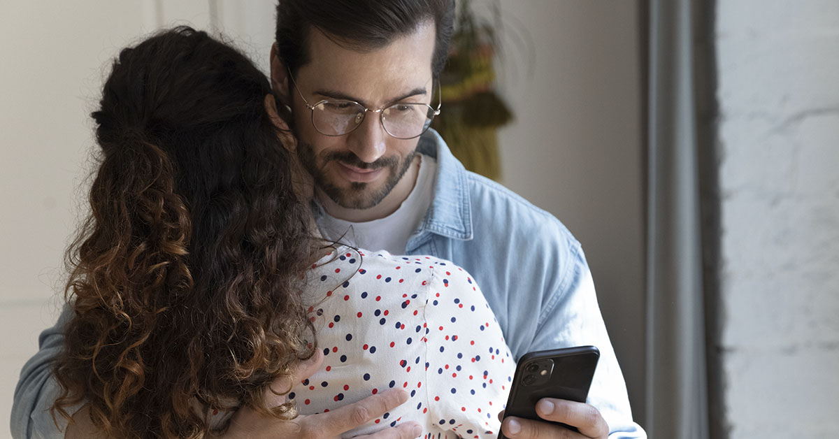 man using smartphone behind partners back while embracing