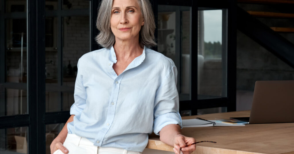 Confident stylish european mature middle aged woman standing at workplace. Stylish older senior businesswoman, 60s gray-haired lady executive leader manager looking at camera in office