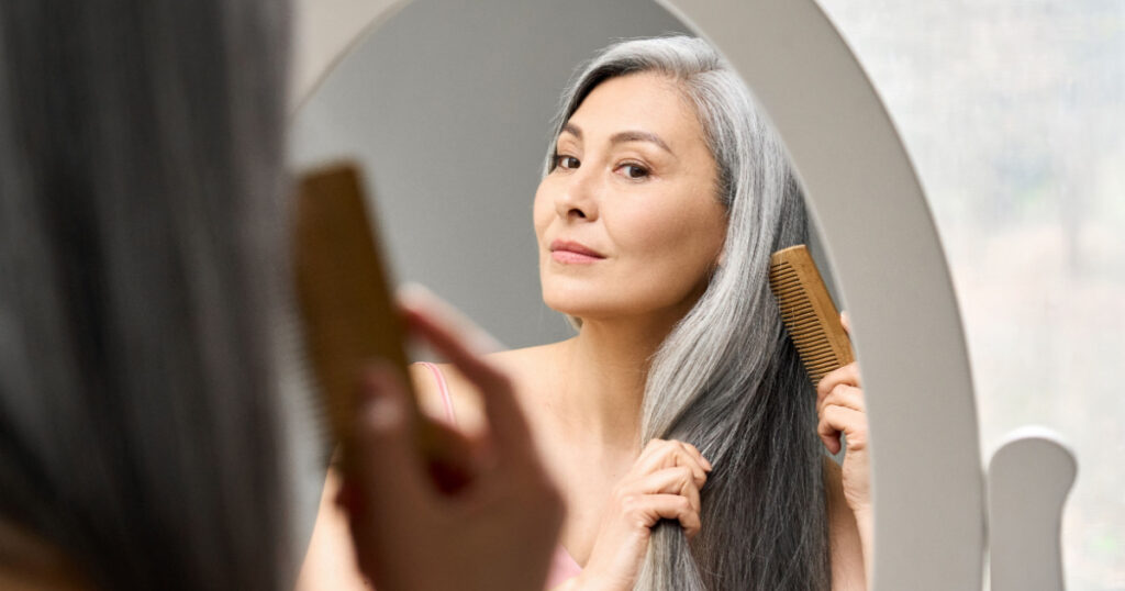 Senior attractive middle 50 years aged asian woman with gray hair looking at mirror reflection combing tangled gray hair