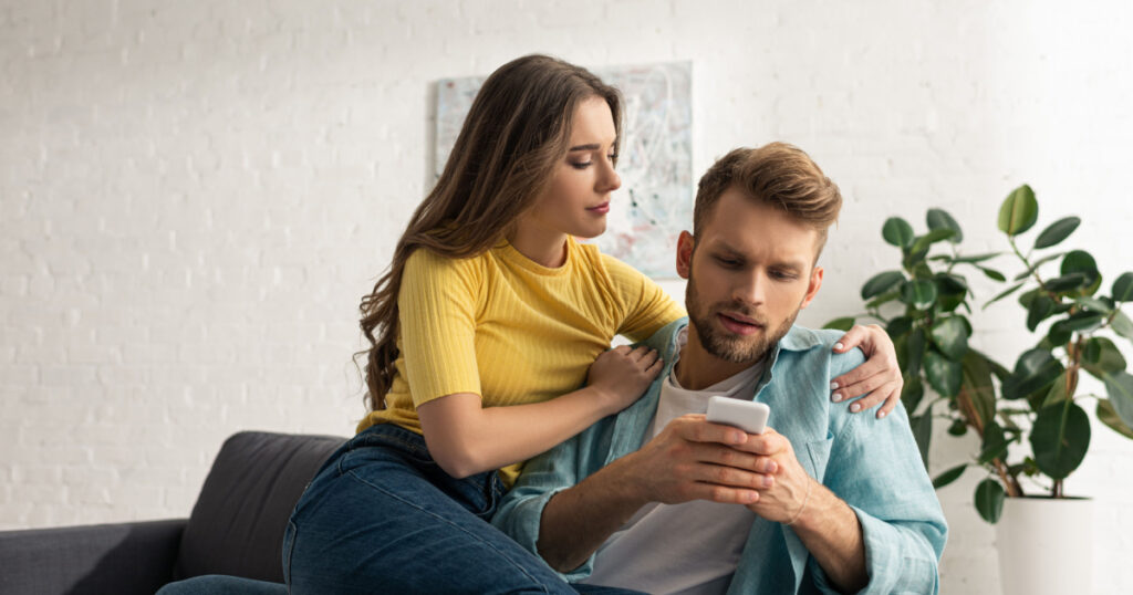 woman embracing addicted boyfriend with smartphone on couch