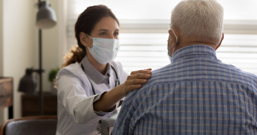 It will be all right. Empathic young female doctor in medical mask on face hold hand on shoulder of elderly man patient encourage to fight with disease. Supportive woman gp comforting old male client