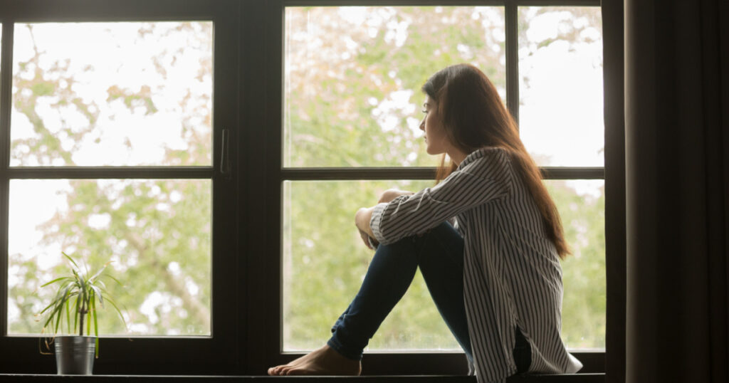 Thoughtful girl sitting on sill embracing knees looking at window, sad depressed teenager spending time alone at home, young upset pensive woman feeling lonely or frustrated thinking about problems