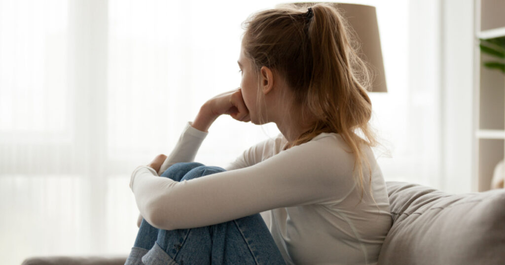 Side view young woman looking away at window sitting on couch at home. Frustrated confused female feels unhappy problem in personal life quarrel break up with boyfriend or unexpected pregnancy concept
