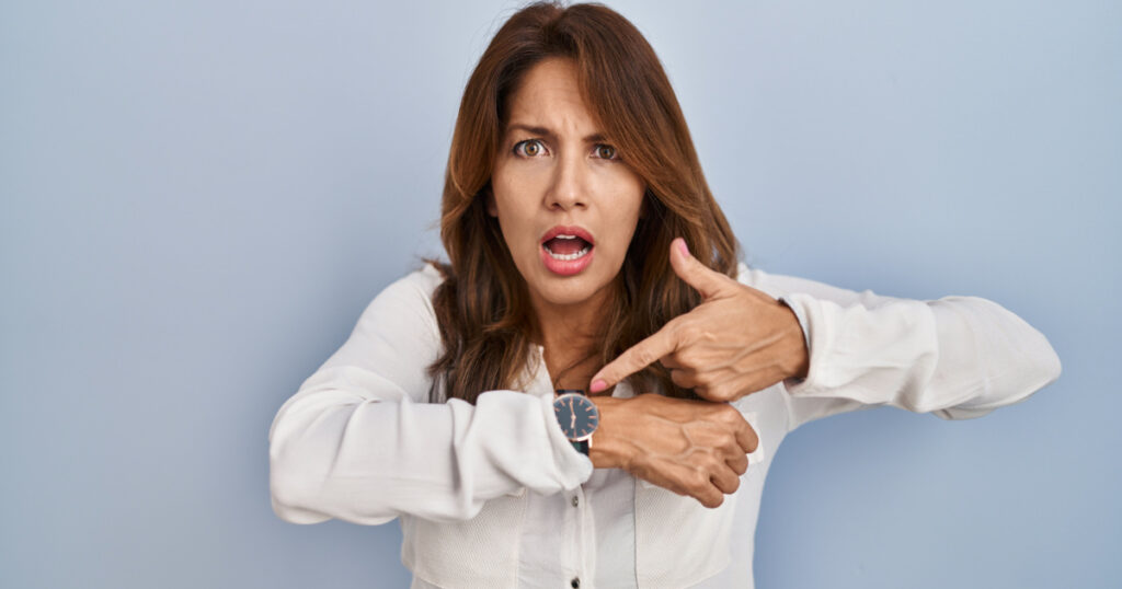 Hispanic woman standing over isolated background in hurry pointing to watch time, impatience, upset and angry for deadline delay 