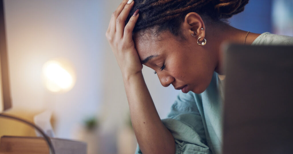 Business woman, depression and stress in an office at night working late on deadline. Tired African entrepreneur person with hands on head for pain, burnout or regret for mistake or fail at work
