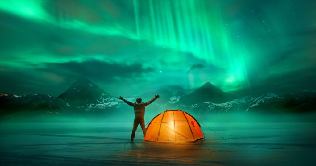 A man camping in wild northern mountains with an illuminated tent viewing a spectacular green northern lights aurora display. Photo composition.
