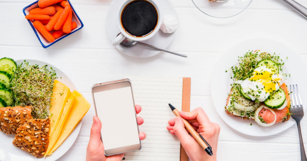 Top view Female hands holding smart phone and writing in notebook on served white wooden table with breakfast dishes. Day diet planning and healthy eating concept. Selective focus, copy space
