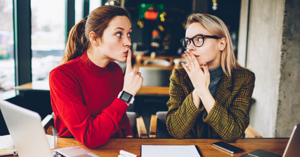 Brunette hipster blogger told secret to wondered colleague and showing sign shh during collaborating at laptop devices in coworking.Excited two best friends gossiping during studying break at netbooks
