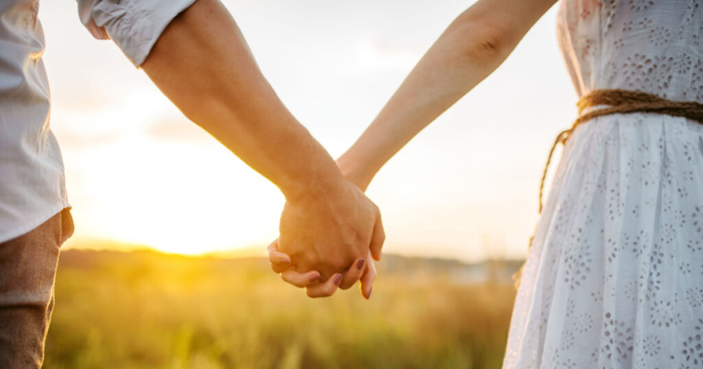 Couple hold hands in green field on sunset
