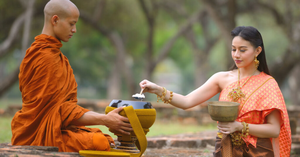 Woman in Thai traditional dress giving food in the bowls of Buddhist monk at Wat Phra Si Sanphet in the early morning, Ayutthaya, Thailand.
