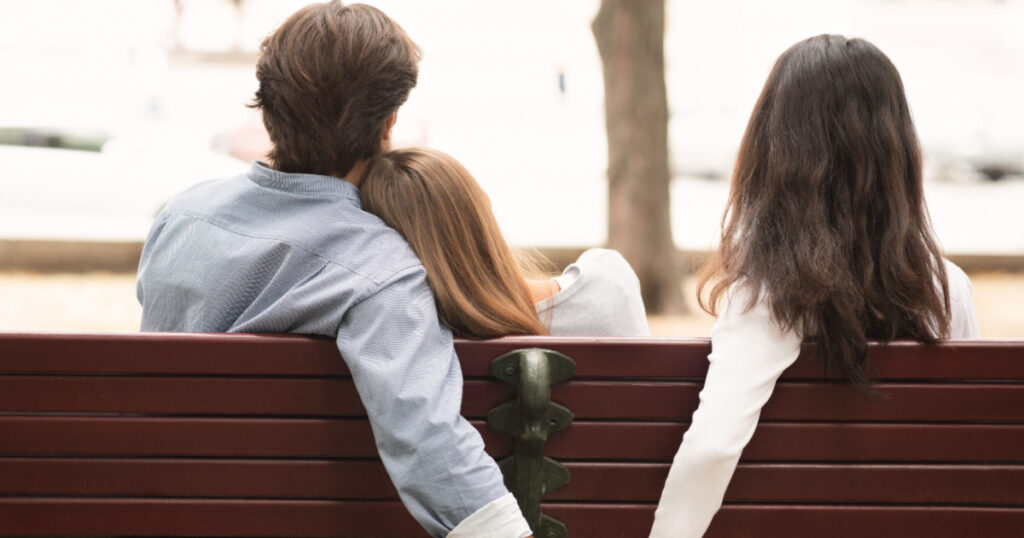 Love Triangle. Cheating Boyfriend Hugging Girlfriend Holding Hands With Her Girl Friend Sitting On Bench Together In Park Outdoor. Back-View
