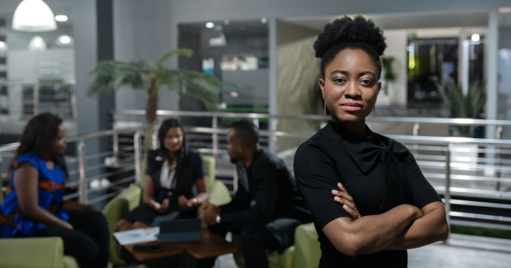 Portrait of a confident young African businesswoman standing with her arms crossed with colleagues meeting in the background

