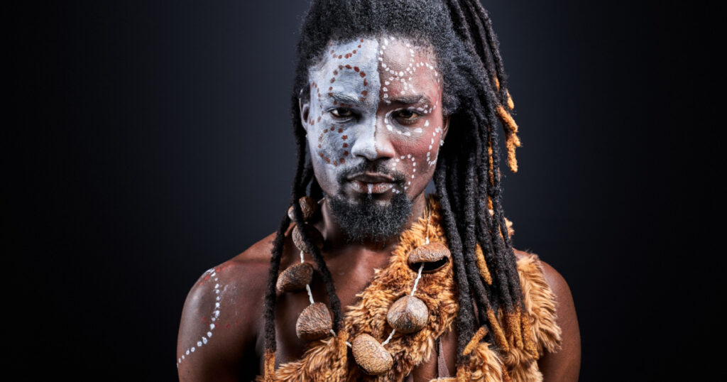 african shirtless guy with colourful make-up isolated on dark studio background, young male tribal has ethnic traditional chain
