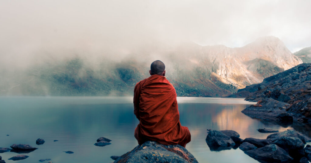 A Tibetan monk from back sitting on the stone near the water in the background of foggy mountains
