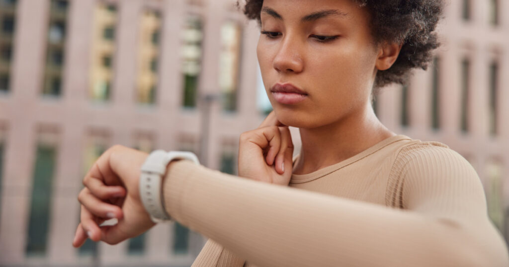 Healthy lifestyle concept. Curly haired woman checks pulse after jogging workout touches neck looks at watch dressed in sportswear stands outdoors against blurred background monitors her health
