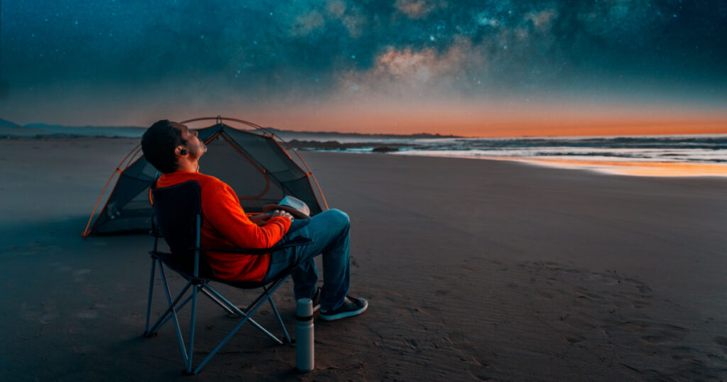 man sitting in a camping chair alone on the beach looking at the starry night next to a tent

