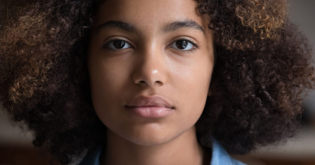 Calm serious beautiful millennial African American woman with thick curly hair looking forward at camera posing indoors. Attractive focused young adult gen z lady face without smile. Close up portrait
