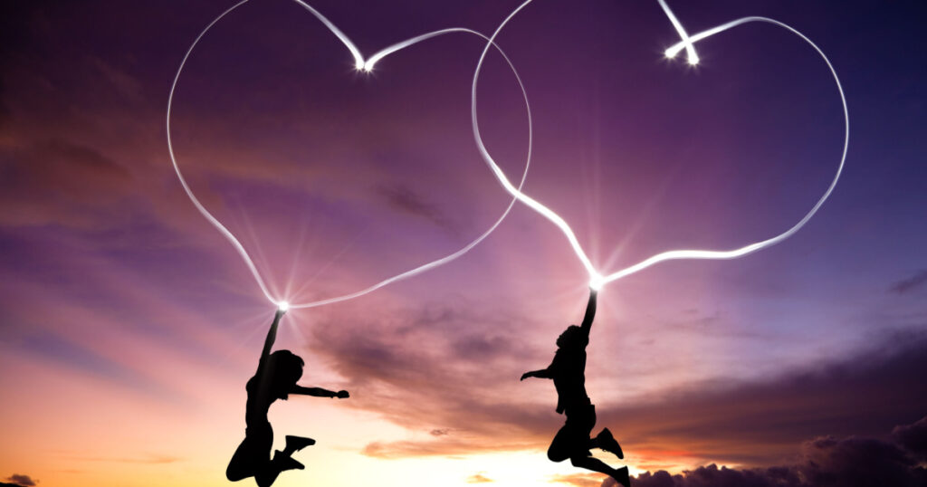 young couple jumping and drawing connected hearts by flashlight in the air on the beach before sunrise
