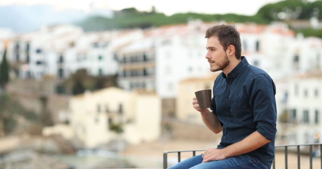Side view portrait of a pensive man drinking coffee and looking away sitting on a ledge in a coast town
