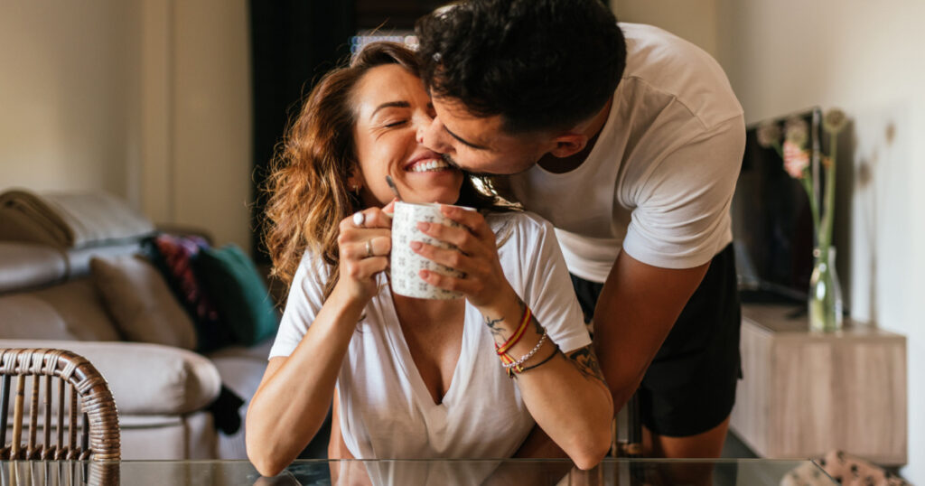 Happy young couple hugging in the living room of their home. Happy couple kissing each other in the face. Family having fun inside a comfortable room having a cup of coffee or tea.
