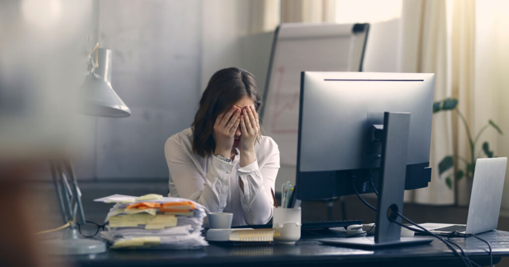 Very stressed business woman and female employee sitting in front of her computer with her hands in front of her eyes, feeling sad and depressed. Too much work - huge pile of paperwork at office table
