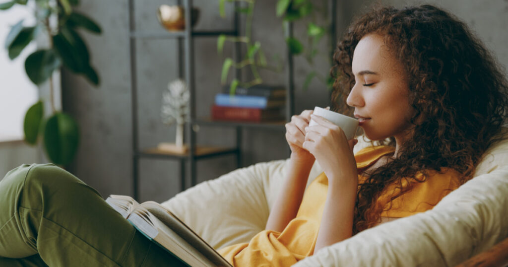 Sideways young woman of African American ethnicity wear t-shirt drink coffee read book close eyes sits in armchair stay at home flat rest relax spend free spare time in living room indoor grey wall
