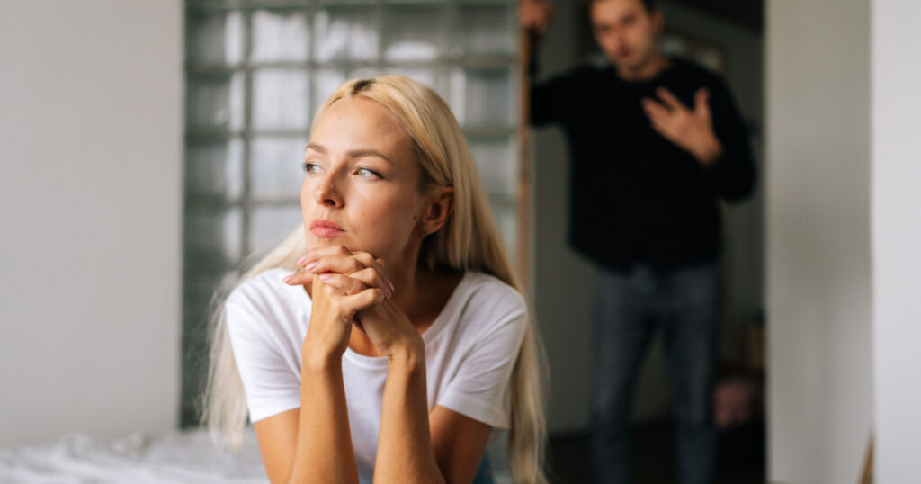 Portrait of tired stressed woman sadness looking away sitting on bed on blurred background of aggressive husband shouting on wife at home