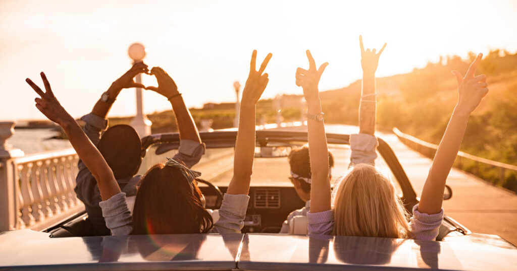 Group of happy young friends in cabriolet with raised hands driving on sunset
