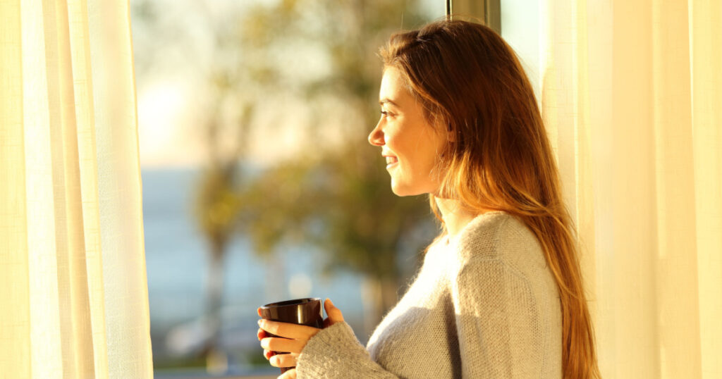 Side view portrait of a single relaxed girl looking the sea outside through a window and holding a coffee mug at sunset in the living room at home
