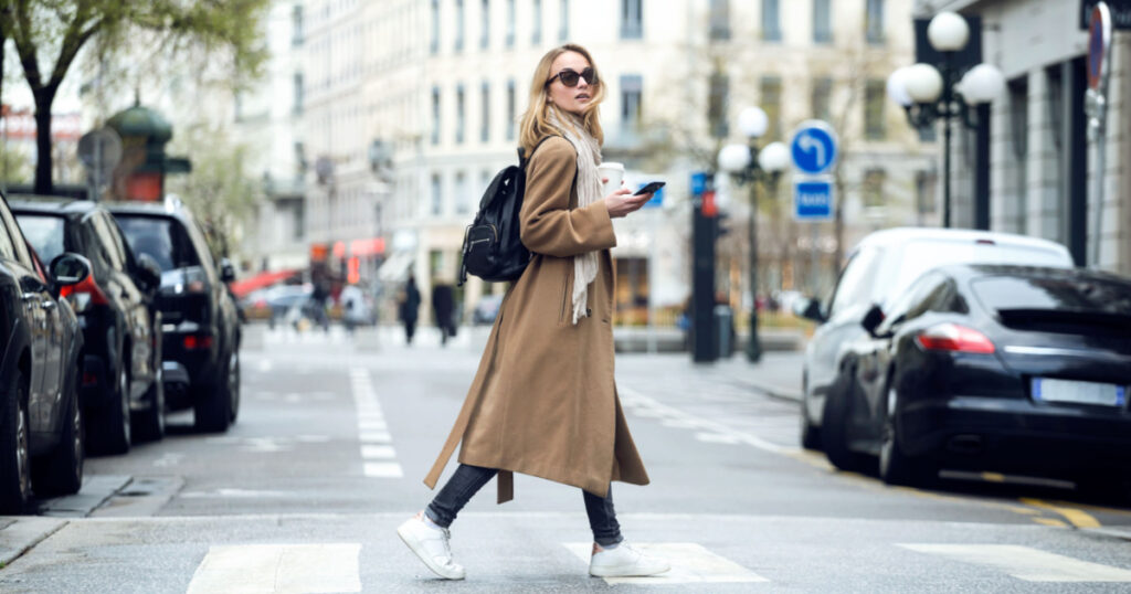 Shot of pretty young woman crossing the street while holding the smartphone and looking sideways.
