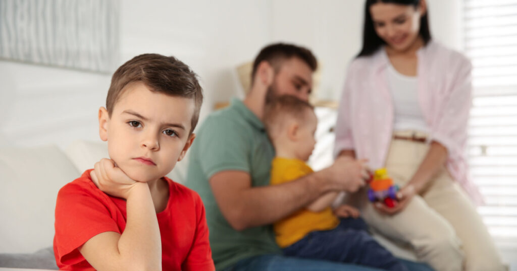 Unhappy little boy feeling jealous while parents spending time with his baby brother at home
