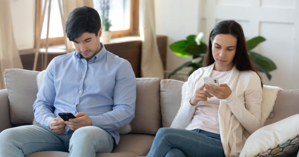 Life online. Concentrated millennial couple internet addiction victims spending leisure time separate sitting on couch at home focused on gadgets mobile phones surfing websites, chatting or shopping
