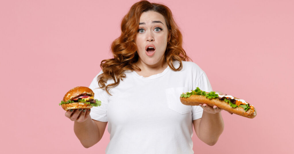 Worried young redhead plus size body positive woman 20s in white casual t-shirt hold american classic fast food hot dog burger looking camera isolated on pastel pink color background studio portrait
