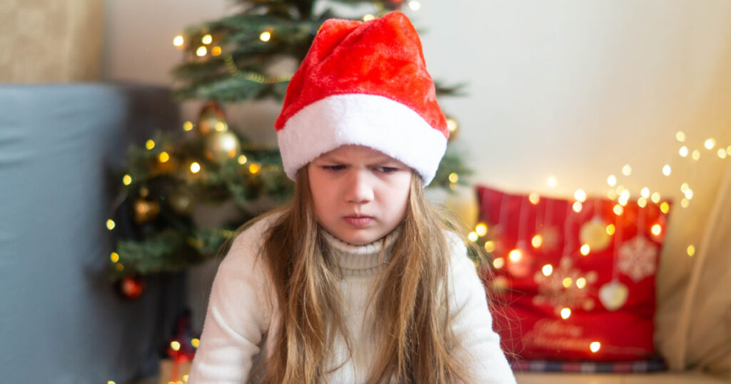 Sad little girl in Santa hat sitting on the background of decorated Christmas tree disappointed with gift. Cute little child feeling unhappy with bad holiday present.
