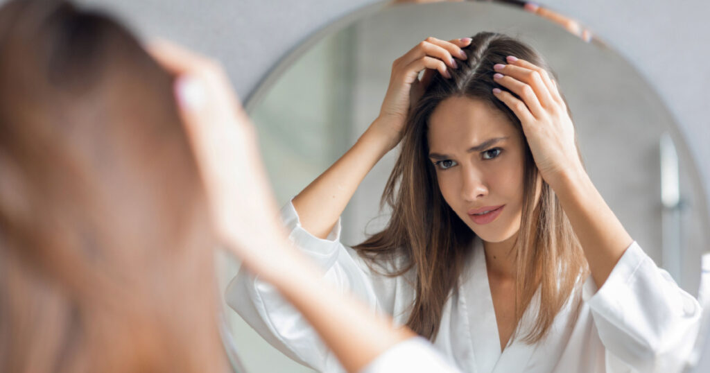 Upset Young Woman Standing Near Mirror In Bathroom And Looking At Her Hair Roots, Frustrated Millennial Lady Suffering Dandruff Or Hairloss Problem, Selective Focus On Reflection, Closeup
