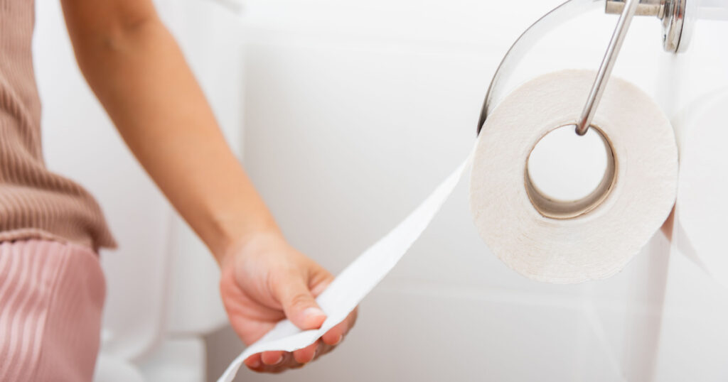 Closeup hand pulling toilet paper roll in holder for wipe, woman sitting on toilet she taking and tearing white tissue on wall to towel clean in bathroom, Healthcare concept
