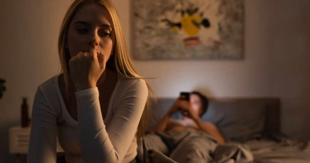 worried woman sitting on bed near blurred boyfriend using smartphone in bedroom
