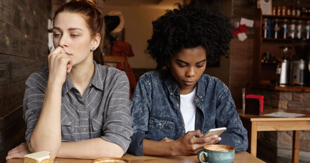 Two unhappy lesbians not talking to each other after having quarrel during lunch at coffee shop: sad redhead woman feeling lonely while her African girlfriend sitting next to her, using mobile phone
