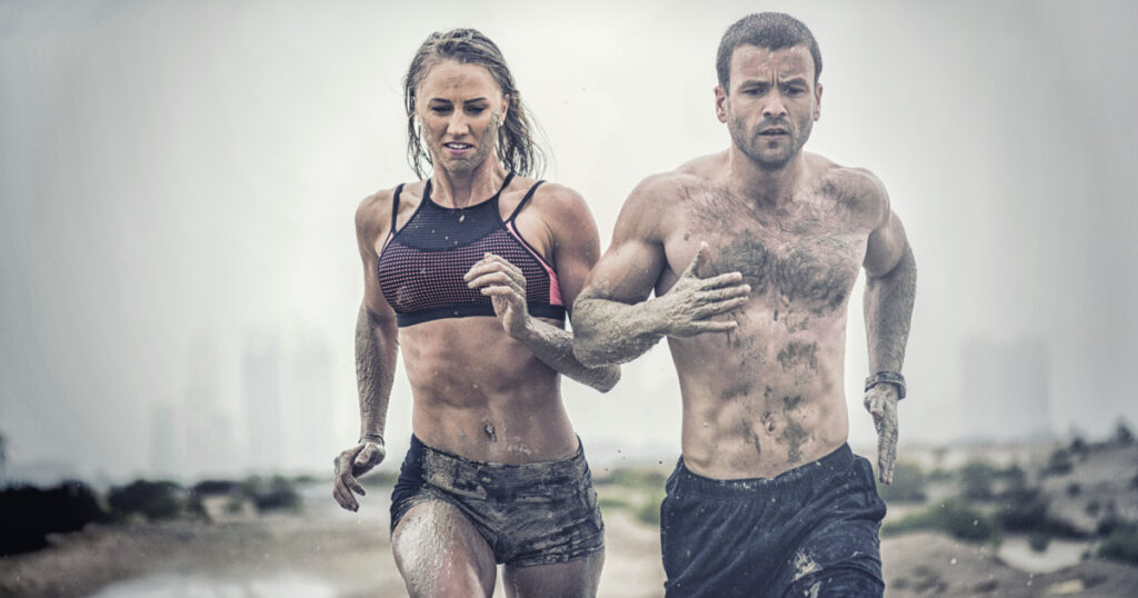 Muscular male and female athlete covered in mud running down a rough terrain with a desert background in an extreme sport race with grungy textured finish
