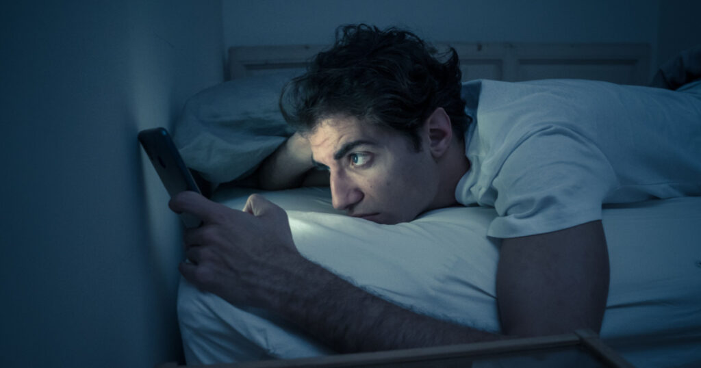 Addicted to social media young man chatting and surfing on the Internet on smart phone at night in bed. Sleepless in dark bedroom with mobile screen light. In insomnia and online network addiction.
