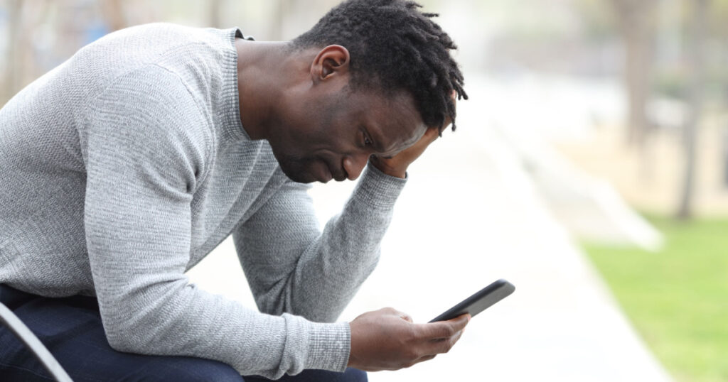 Side view portrait of a sad black man complaining checking mobile phone sitting on a bench in a park
