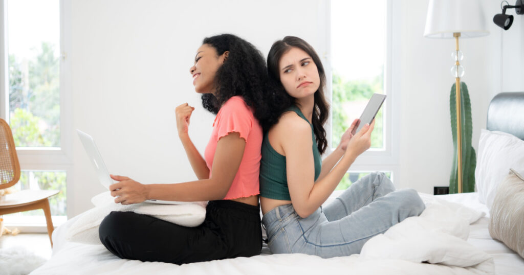 Two young women sitting on bed in bedroom and using laptop and tablet for play the game.
