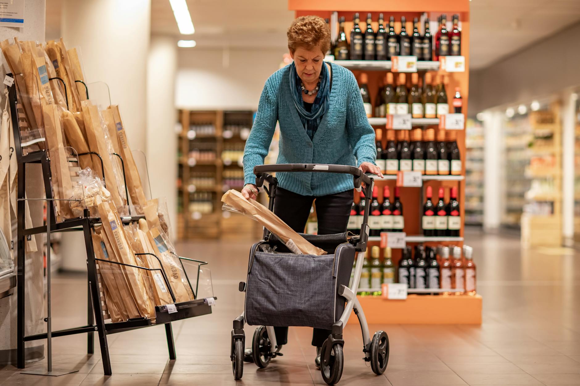 Elderly woman using a rollator walker while shopping for bread in a supermarket.