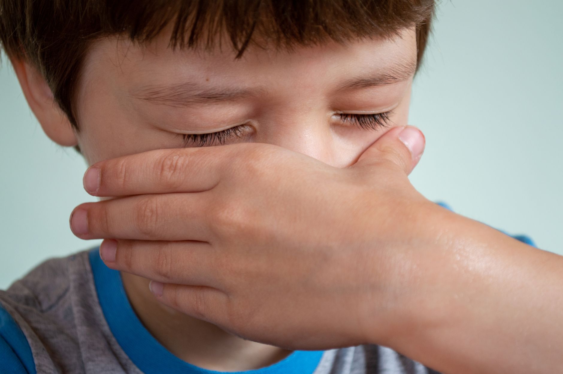 Crop little boy with closed eyes and brown hair crying and wiping tears from cheeks against white background