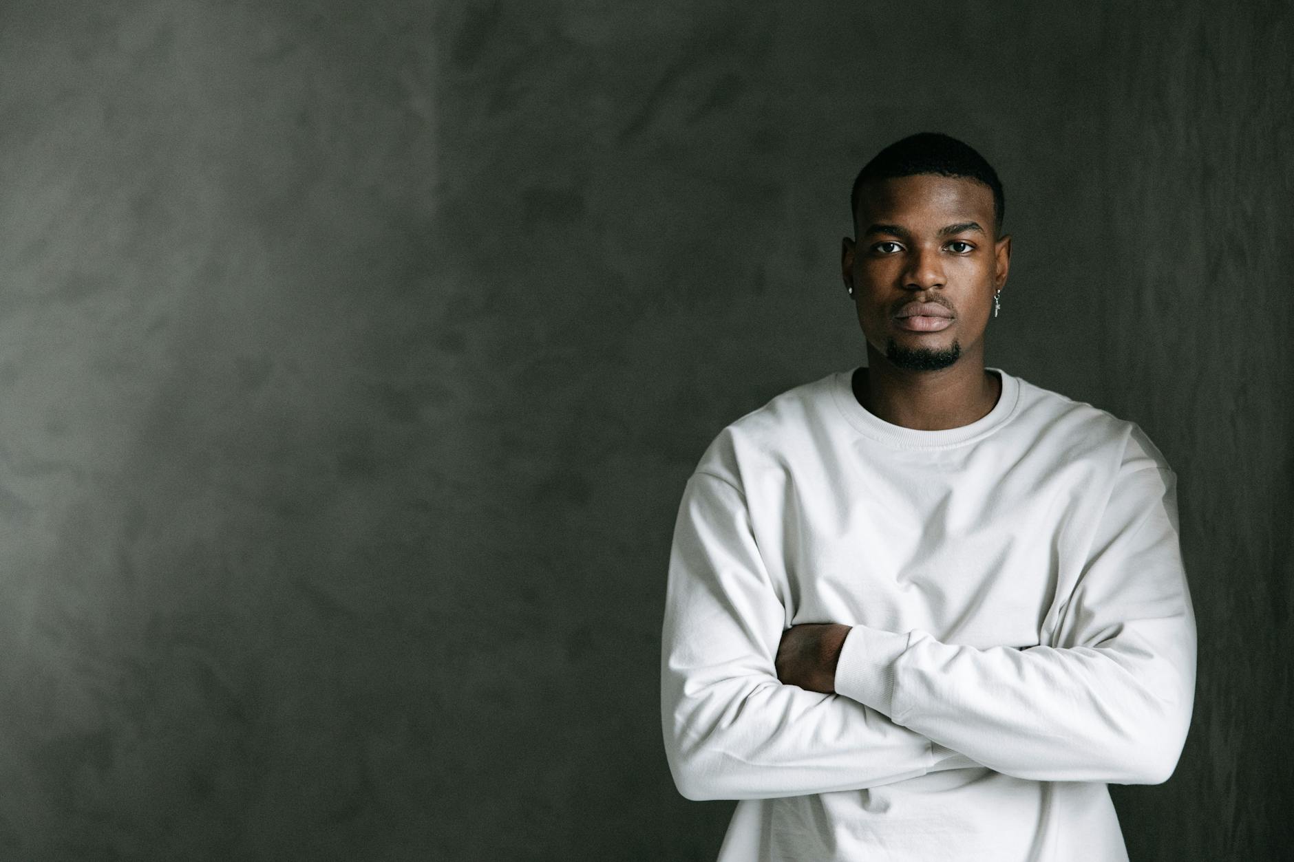 Portrait of a serious man in a white shirt with arms crossed, standing against a textured gray background.